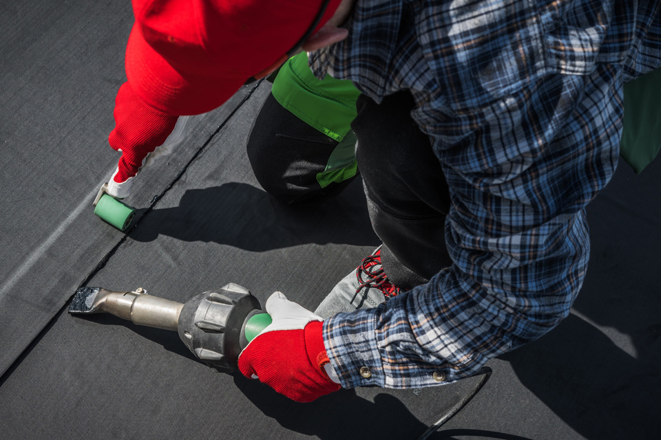 Overhead view of plumber in red gloves and blue plaid shirt holding pipe wrench and metal coupling on grey floor.