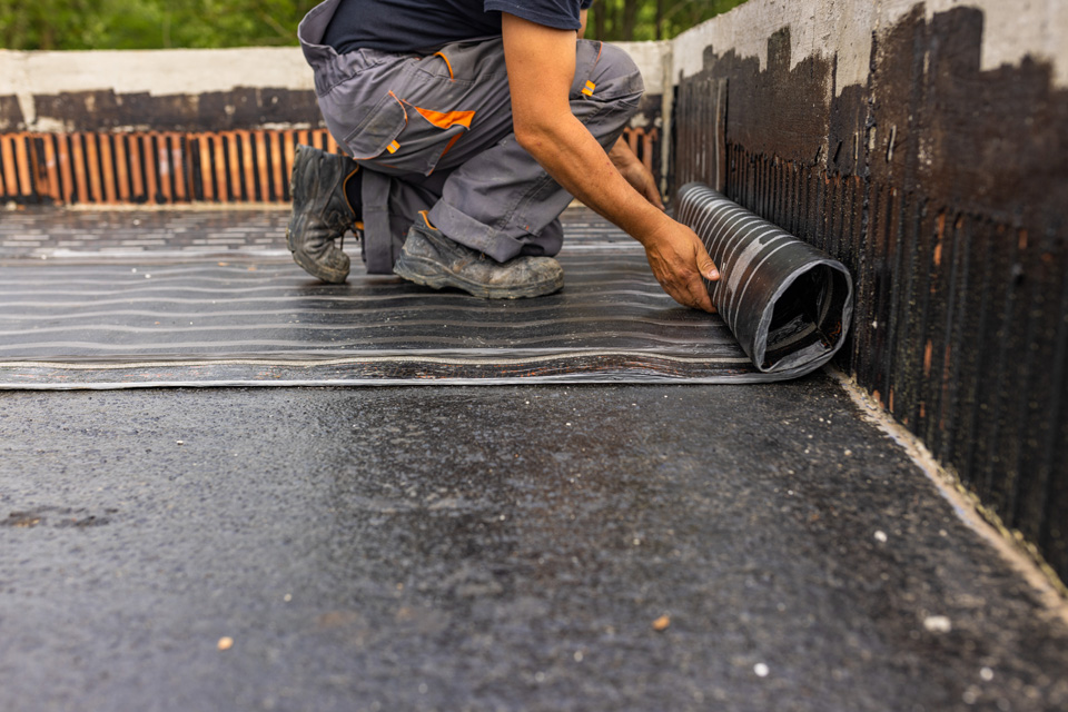 Worker installing waterproof membrane on flat roof with rolled material and tools on concrete surface.