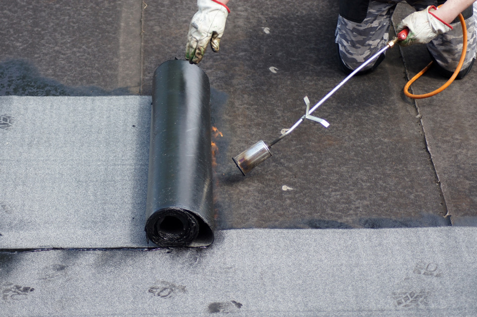 Worker applying bitumen membrane to flat roof during installation with heat gun and protective gloves.