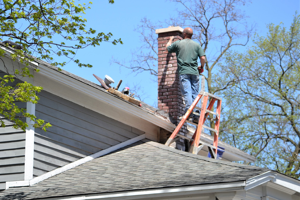 Roofer on ladder installing shingles on residential roof with brick chimney on clear sunny day.