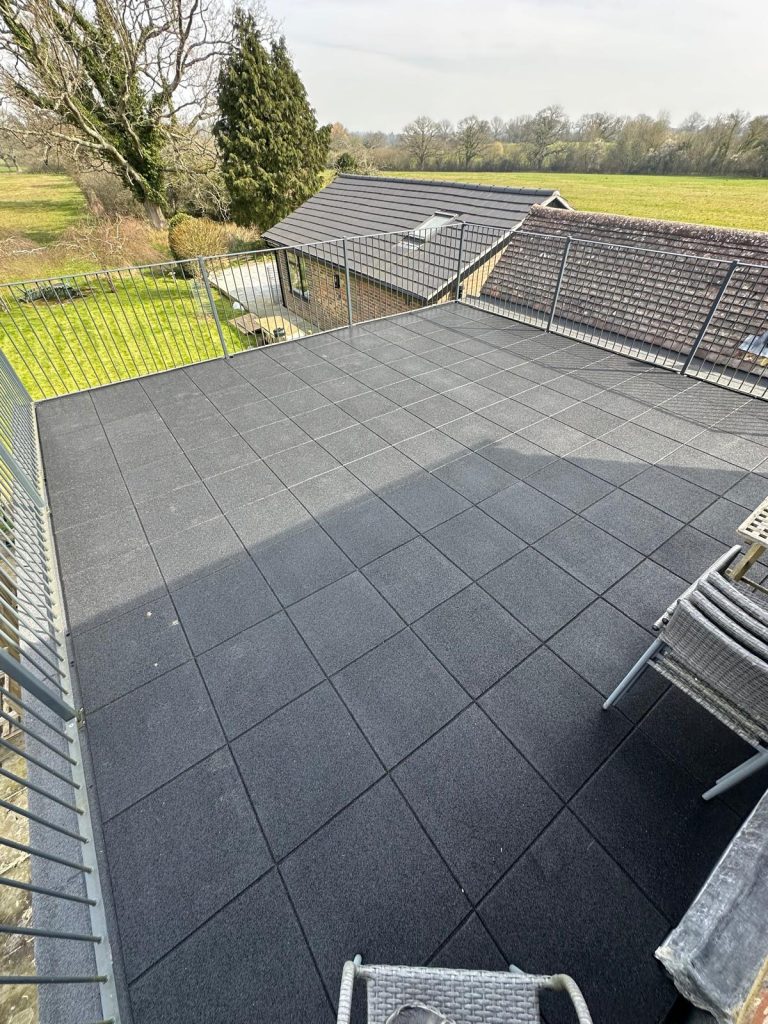 Modern rooftop terrace with dark gray tiles, metal railing, and countryside views overlooking green fields and trees