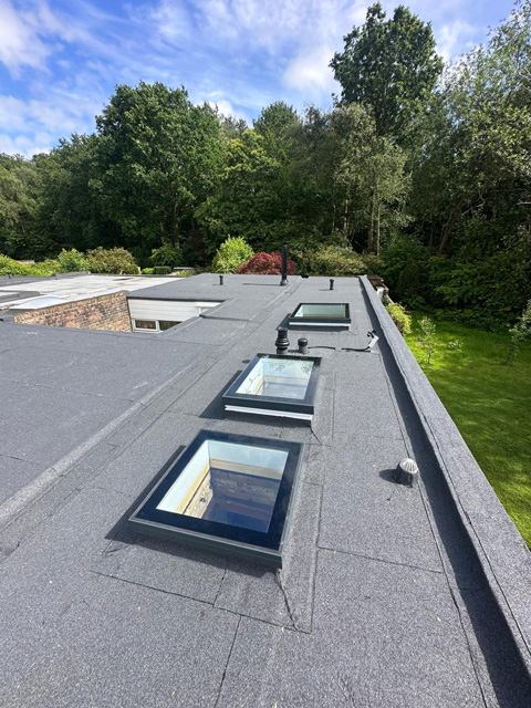 Modern flat roof with multiple skylight windows and grey membrane covering, surrounded by green trees and manicured garden.